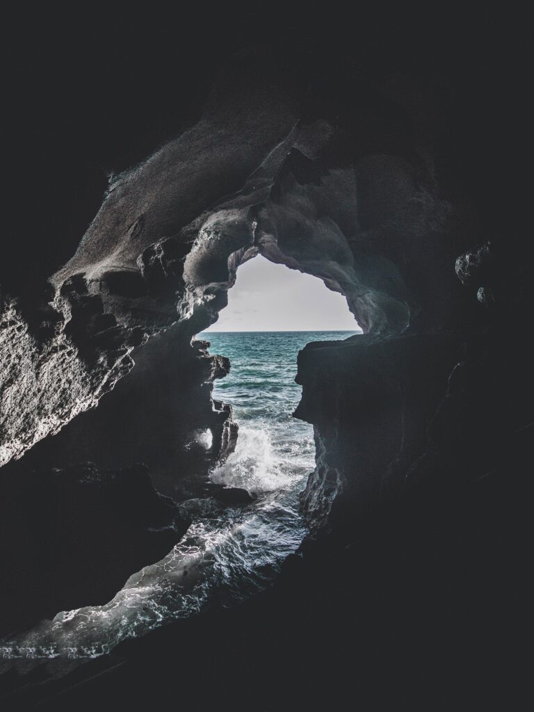 Dramatic sea cave interior with jagged rocks framing vibrant blue ocean waves crashing sunlight Wallpaper