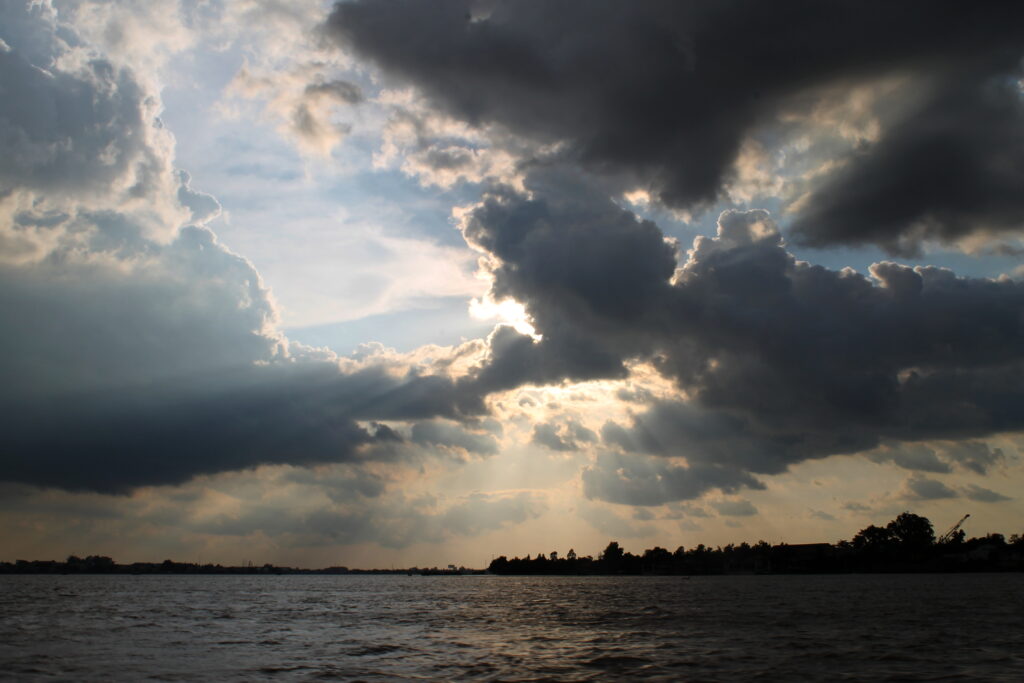 Dramatic sea landscape with dark clouds breaking sunlight over water and distant tree silhouettes Wallpaper