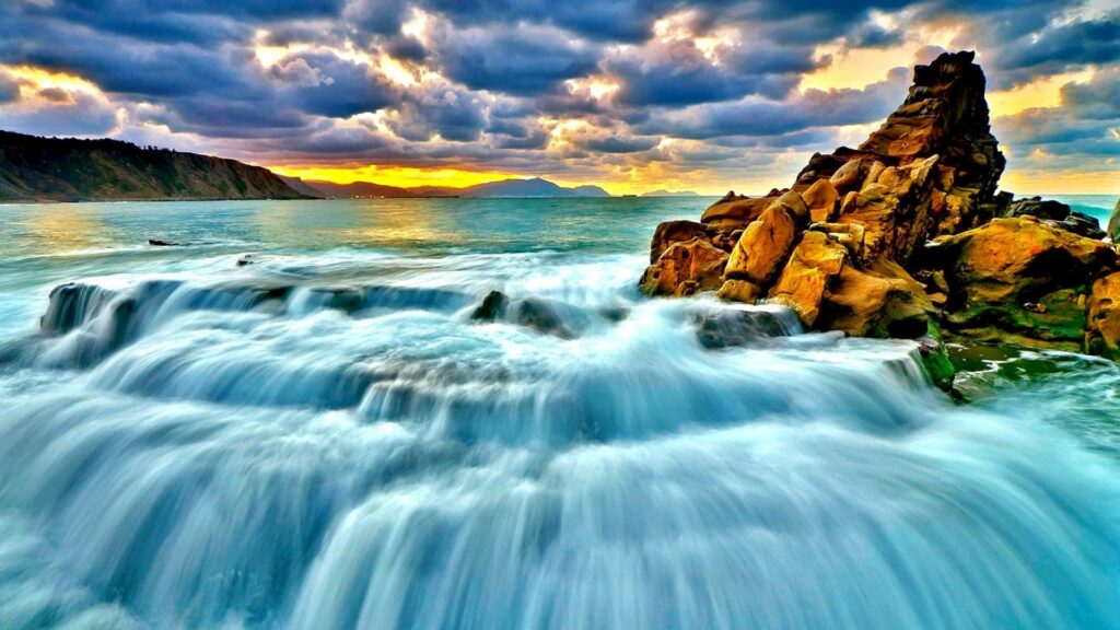 Dramatic seascape with powerful waves over rugged rocks at sunset vibrant golden blue sky wallpaper