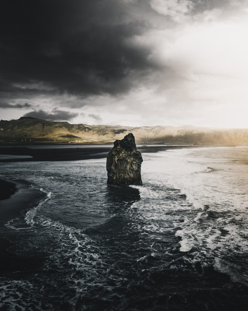 Rugged rock formation on windswept coast with crashing waves and moody overcast sky wallpaper