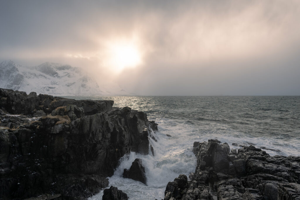 Dramatic seascape with rugged rocks foamy waves misty mountains and soft sunlight breaking clouds Wallpaper