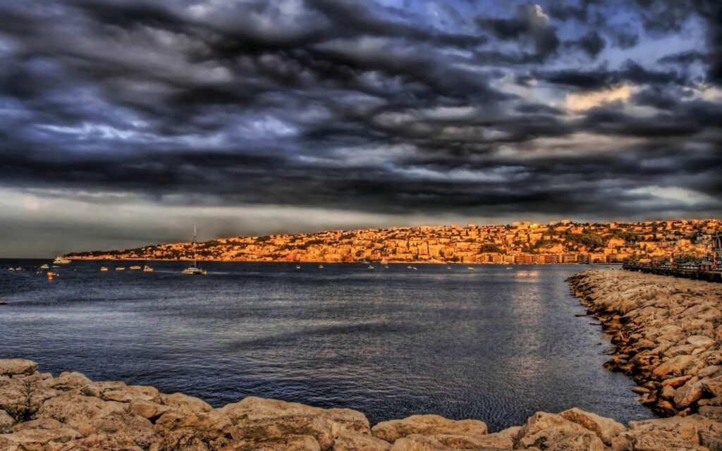 Dramatic seascape with stone pier calm blue waters moody clouds and golden sunlight coastal city Wallpaper