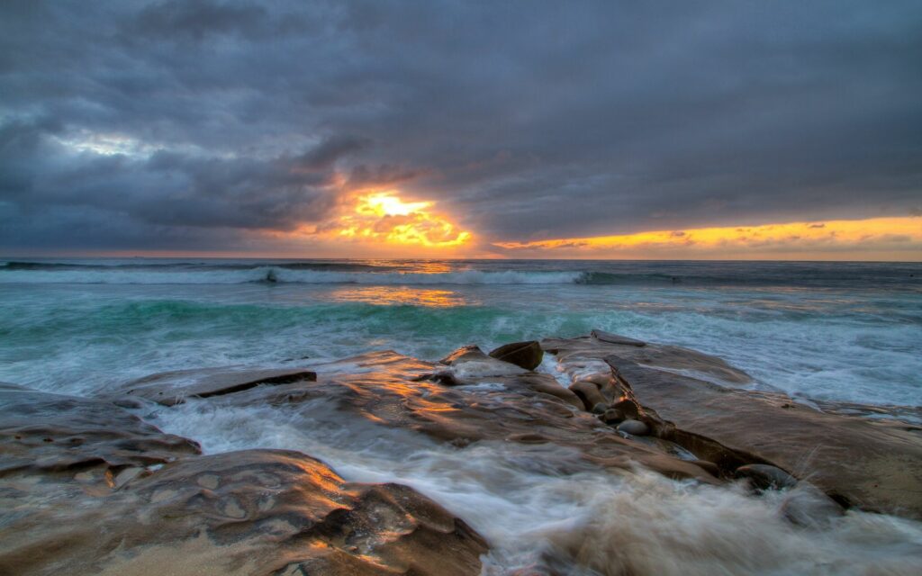 Dramatic seascape with textured rocks waves and vibrant orange sunset reflecting on ocean surface Wallpaper