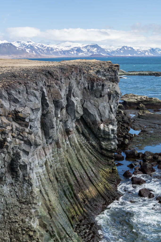 Dramatic seaside cliff with rugged rock formations and crashing waves by snowy mountains Wallpaper