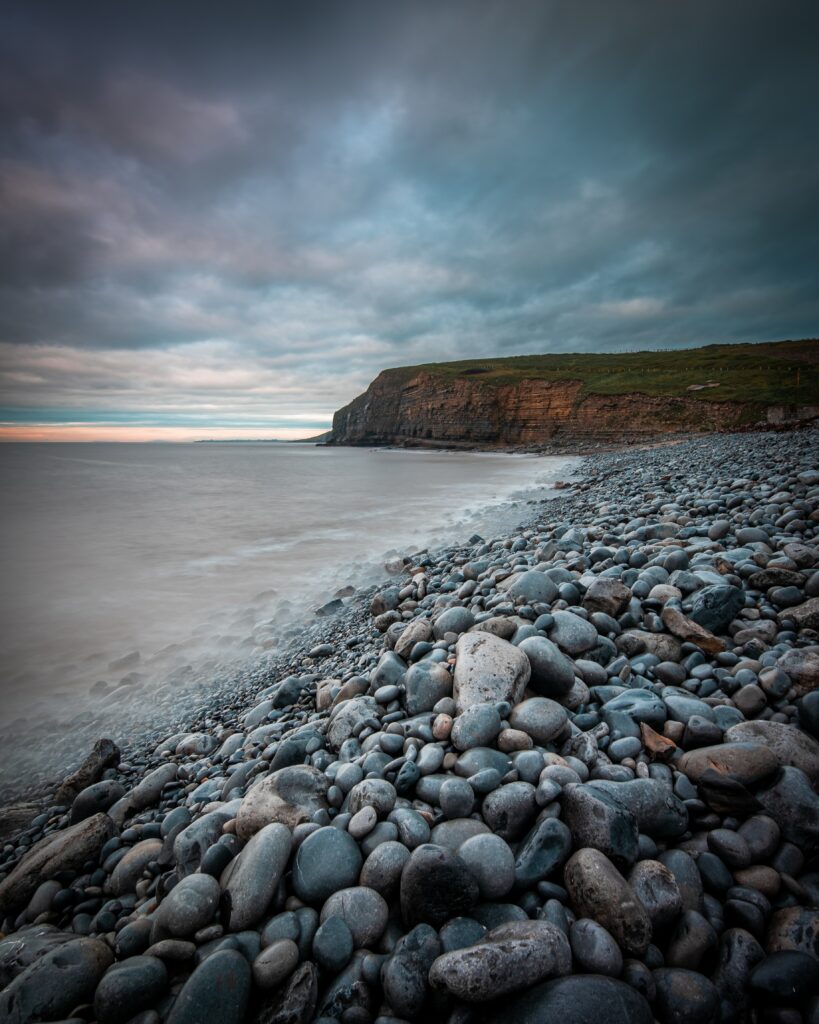 Dramatic seaside wallpaper with smooth pebbles calm sea moody sky and rugged cliffs background