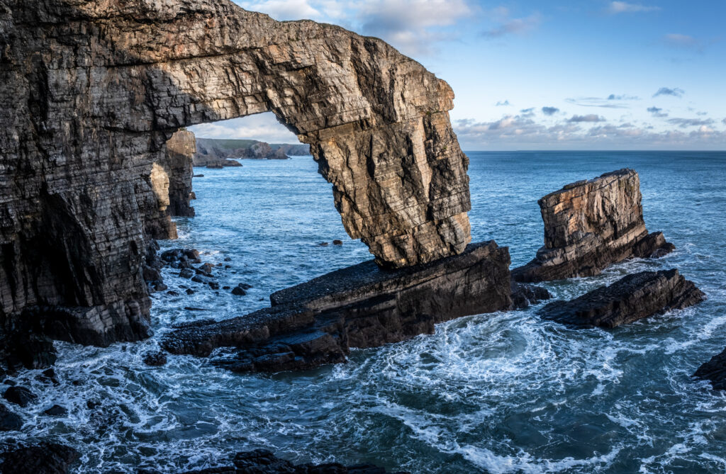 Dramatic seaside landscape with massive natural rock arch and swirling blue waves under clear sky Wallpaper