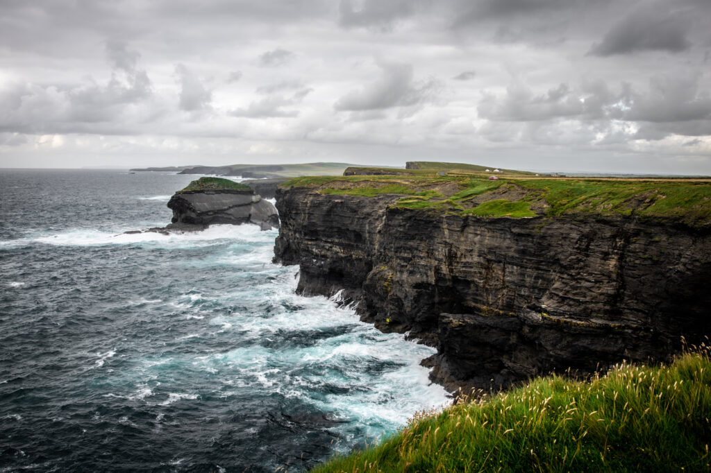 Dramatic seaside landscape with powerful waves crashing rugged rocks and green cliffs under moody sky Wallpaper