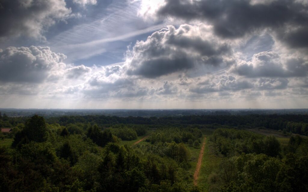 Dramatic cloudy sky casting shadows over lush green trees and winding roads in countryside Wallpaper