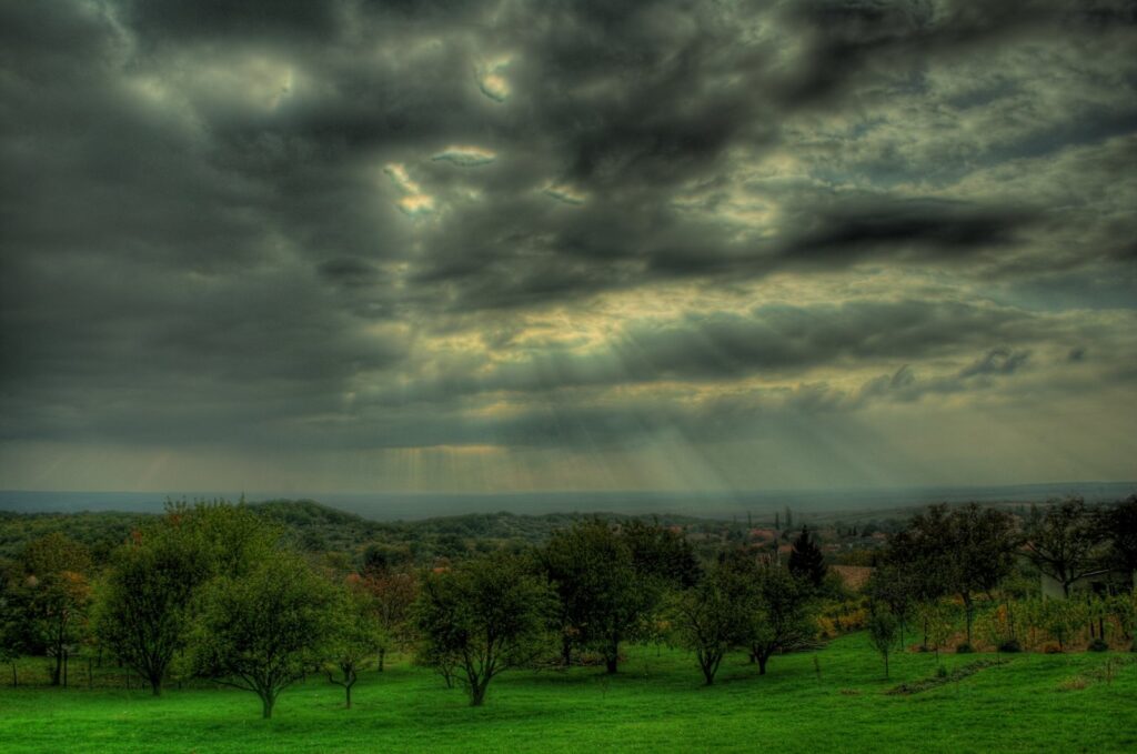 Dramatic cloudy sky with sun rays over lush green landscape and scattered trees wallpaper background