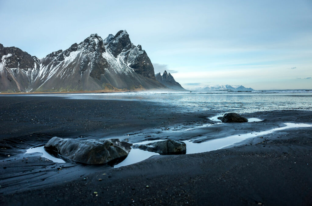 Snow-capped mountains above rugged black sand coastline with tidal pools under overcast sky wallpaper
