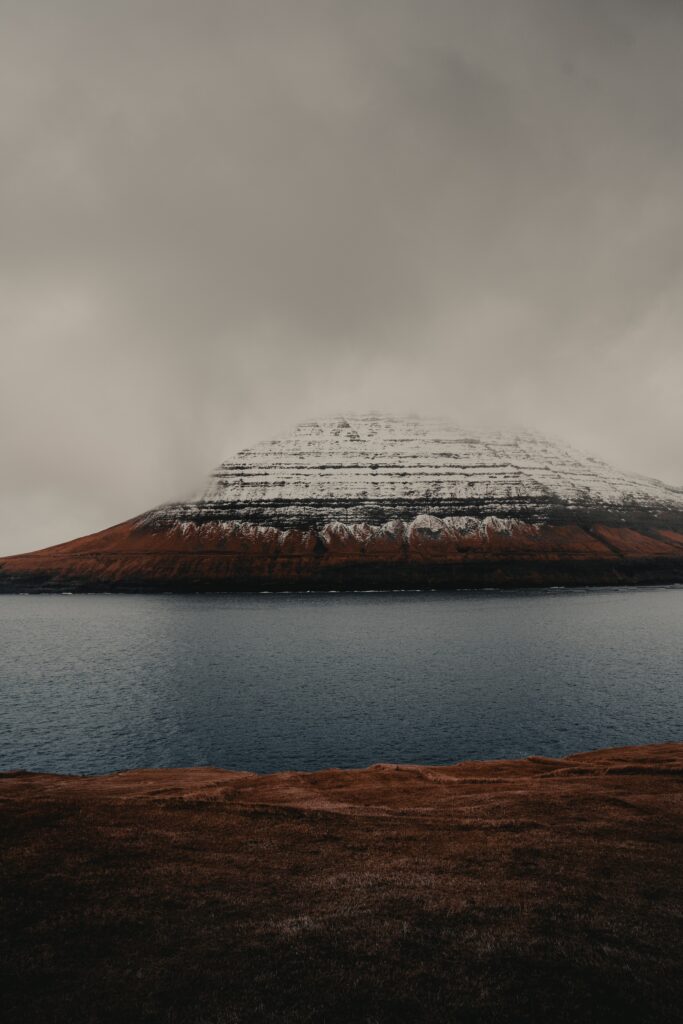 Snow-capped rocky mountain above calm sea with reddish grass and cloudy moody sky wallpaper