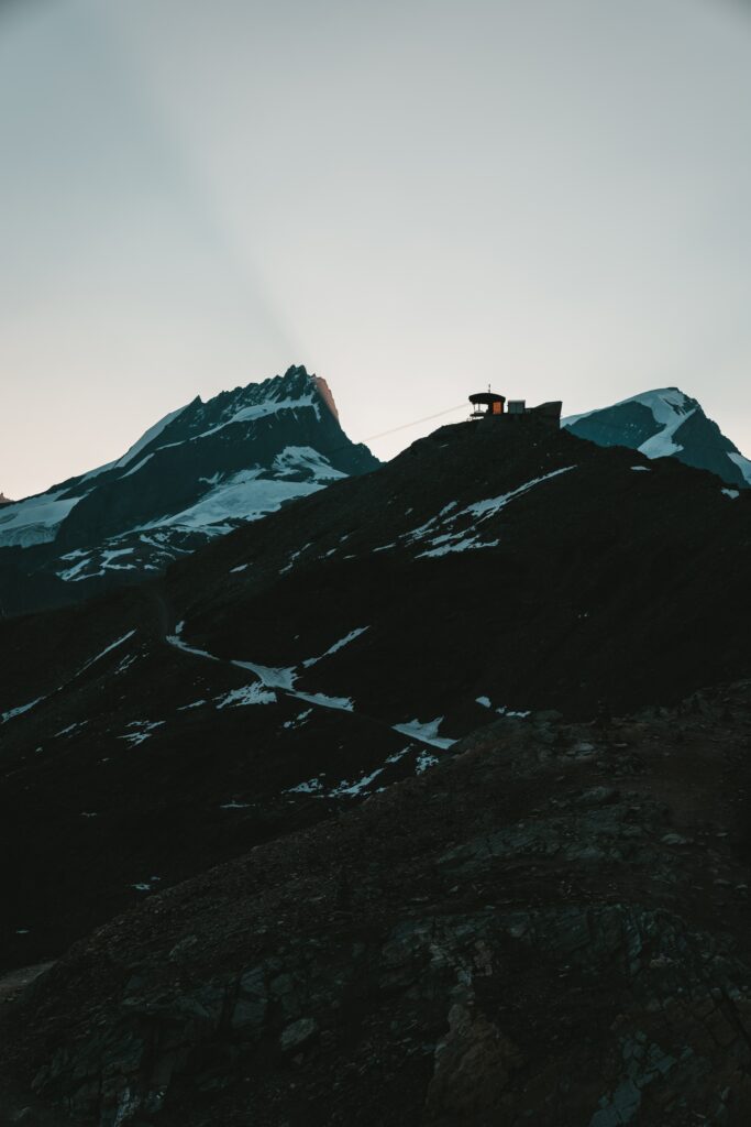 Dramatic snow-dusted rocky mountain peaks at dawn with silhouetted structures in soft sky background Wallpaper