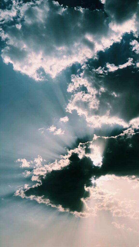Dramatic clouds with radiant sun rays piercing through blue sky creating stunning light contrast Wallpaper