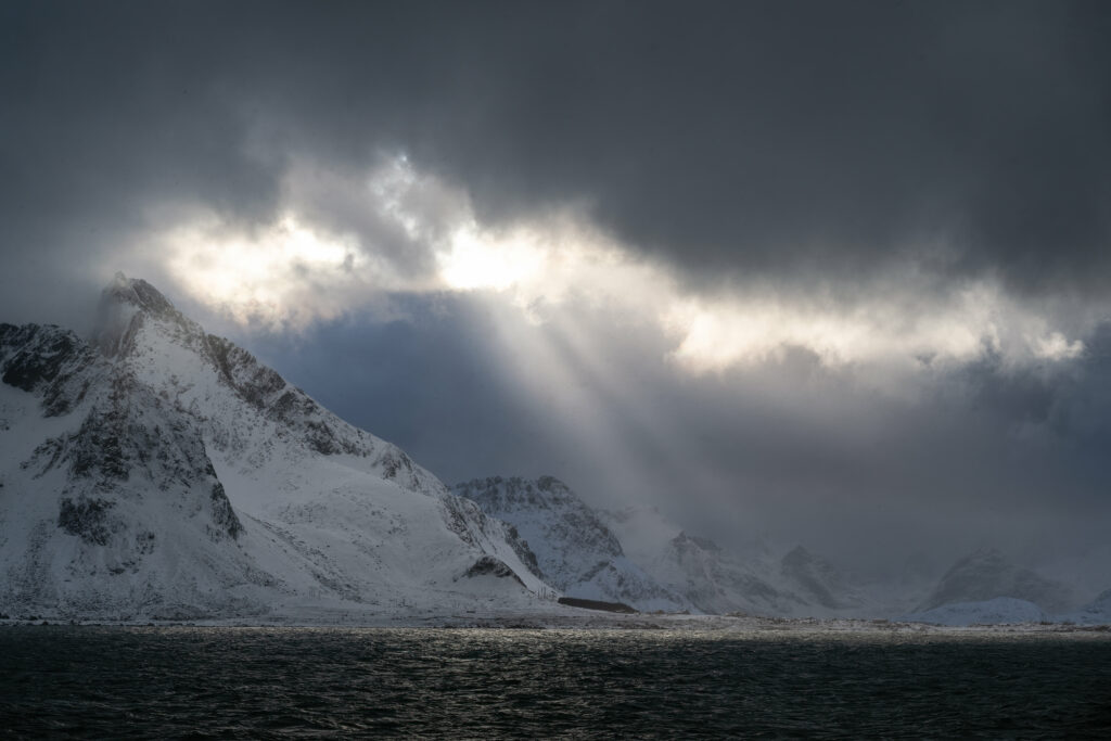 Sunlight breaks through clouds over snowy mountains casting silvery glow on dark rippling sea Wallpaper