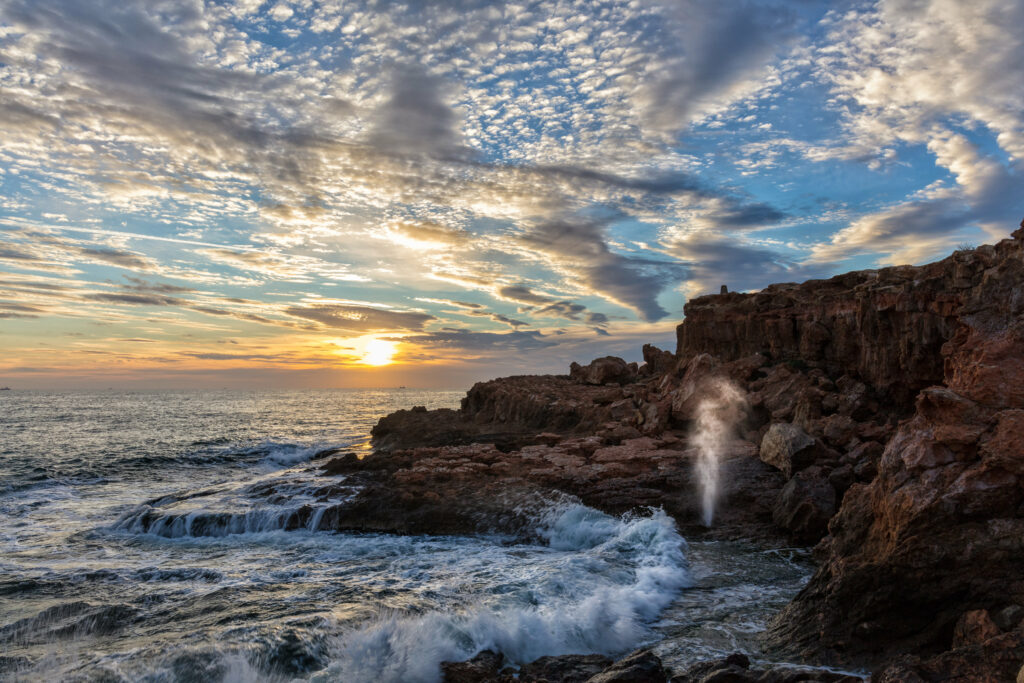 Dramatic coastal sunrise with crashing waves and rugged rocks under vibrant golden clouds Wallpaper