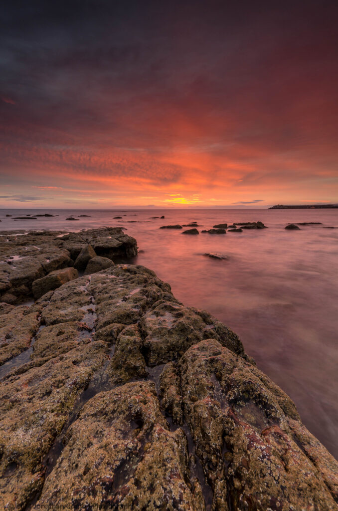 Dramatic sunset over tranquil sea with golden purple sky and rugged stones coastal wallpaper background