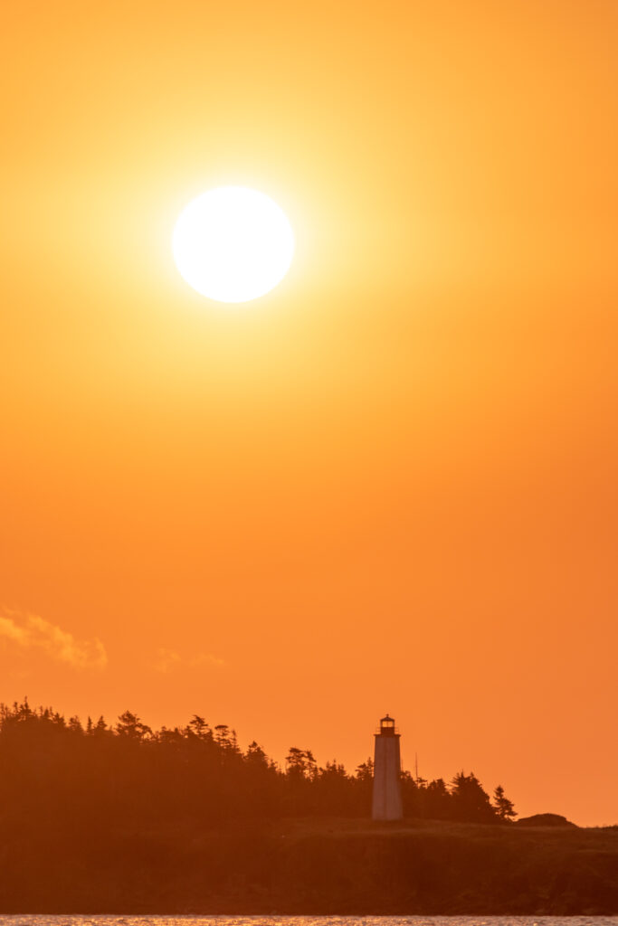 Dramatic sunset over tranquil shore with glowing sun lighthouse and tree silhouettes at dusk Wallpaper