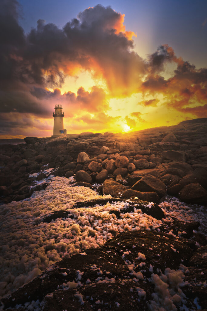 Lighthouse on rocky shore at sunset with golden light dark clouds and foamy surf landscape wallpaper