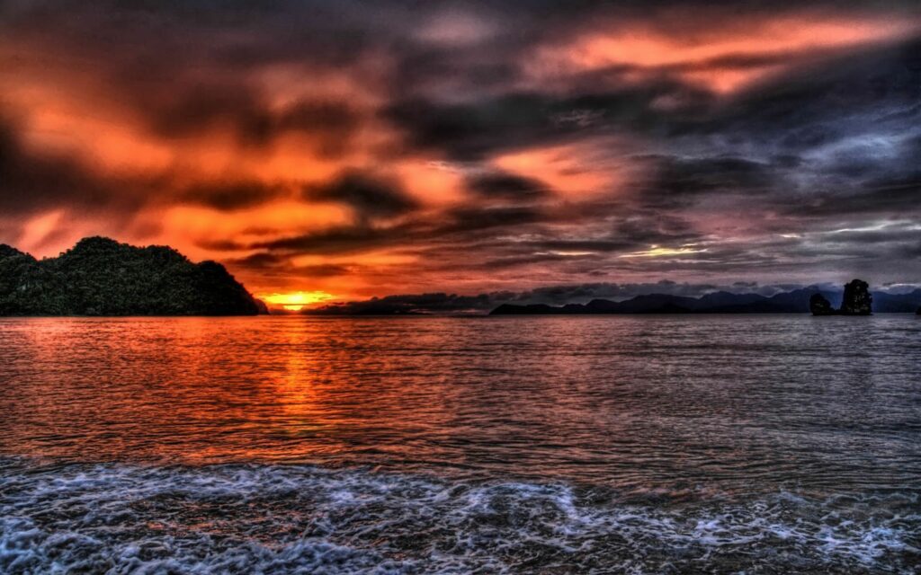 Dramatic sunset with orange red sky reflecting on calm sea and mountain silhouettes at horizon Wallpaper