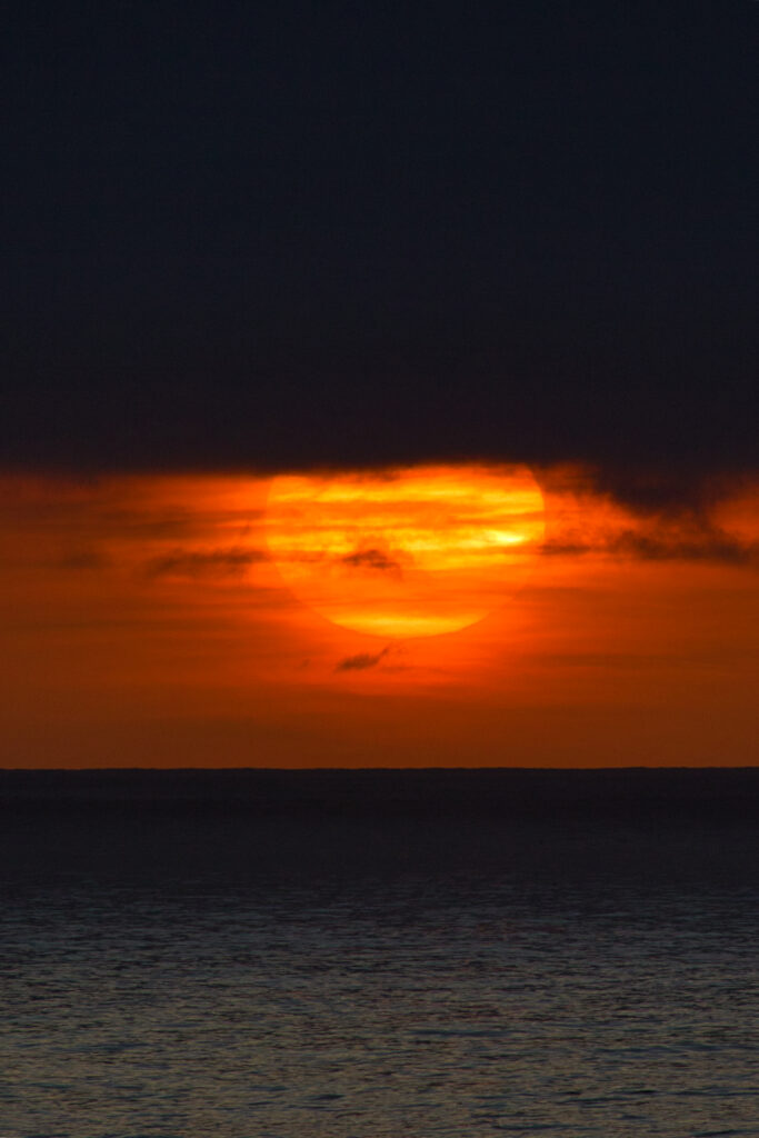 Sunset over calm sea with glowing orange sun behind dark clouds and vibrant reflections wallpaper