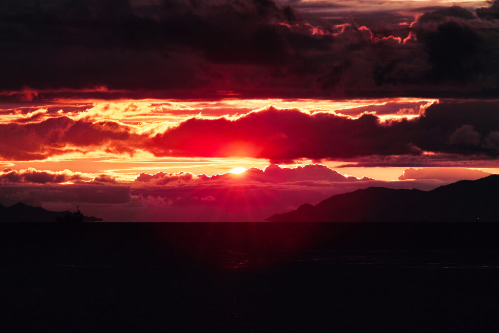 Dramatic sunset with vibrant red and orange clouds over silhouetted mountains and calm sea wallpaper