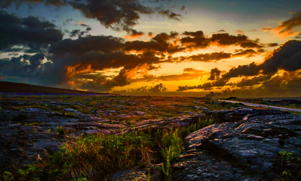 Dramatic sunset over grassy field with scattered stones and textured clouds in rocky terrain Wallpaper