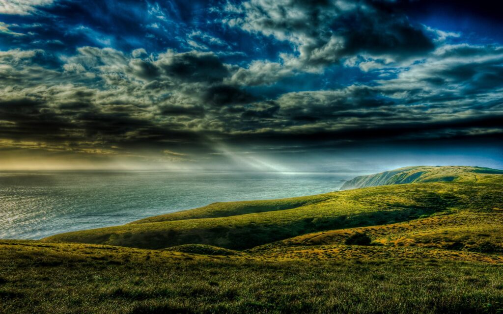 Dramatic sunset over green hills and calm sea with golden light beams breaking clouds wallpaper