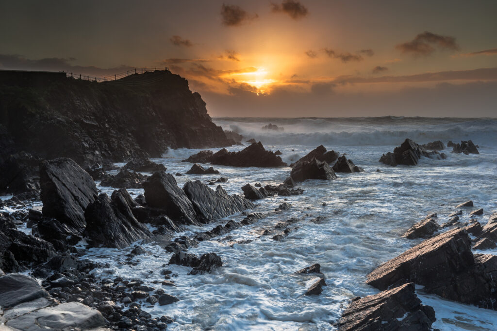 Dramatic sunset over restless sea with vibrant orange gold hues jagged rocks and crashing waves Wallpaper