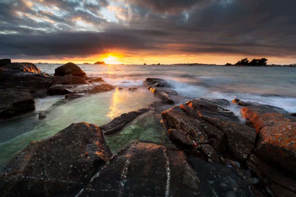 Dramatic sunset over rugged sea rocks with shimmering tide-washed stones coastal landscape Wallpaper