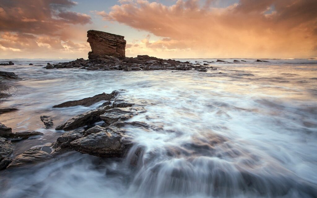Dramatic seascape with waves over dark stones and glowing sunset sky with distant rock formation Wallpaper