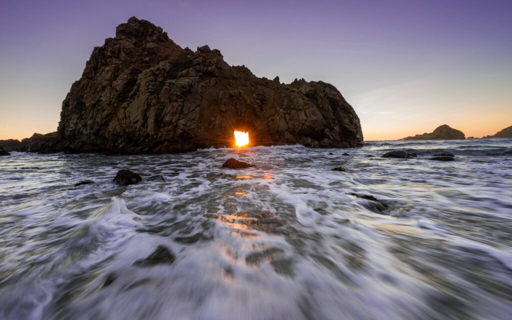 Sunset seascape with ocean tide crashing dark rocks under natural cliff arch coastal wallpaper