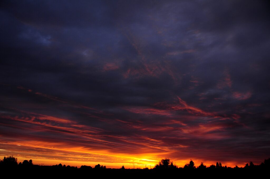 Dramatic sunset with deep orange and red hues silhouetted trees under dark swirling clouds Wallpaper