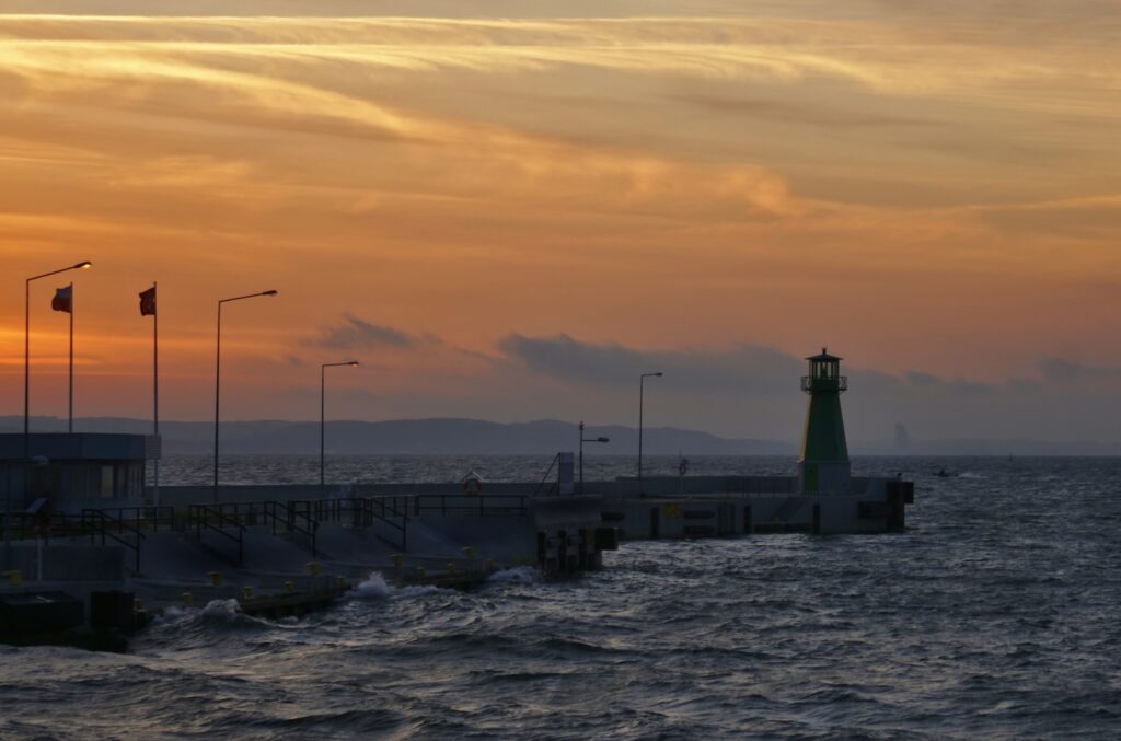 Dramatic sunset over pier with vibrant green lighthouse and rolling sea waves coastal wallpaper