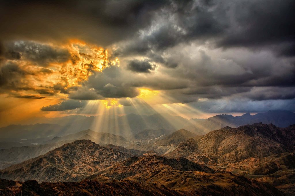 Dramatic mountain range under stormy clouds with golden sunshine rays illuminating terrain Wallpaper