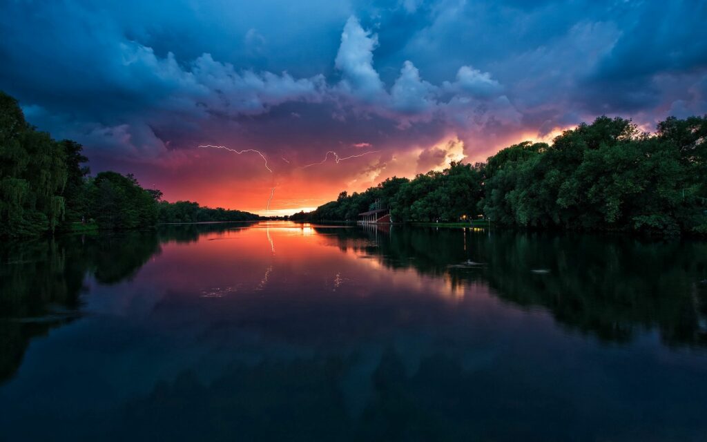Dramatic thunderstorm with vivid lightning over calm river sunset lush green trees reflection wallpaper