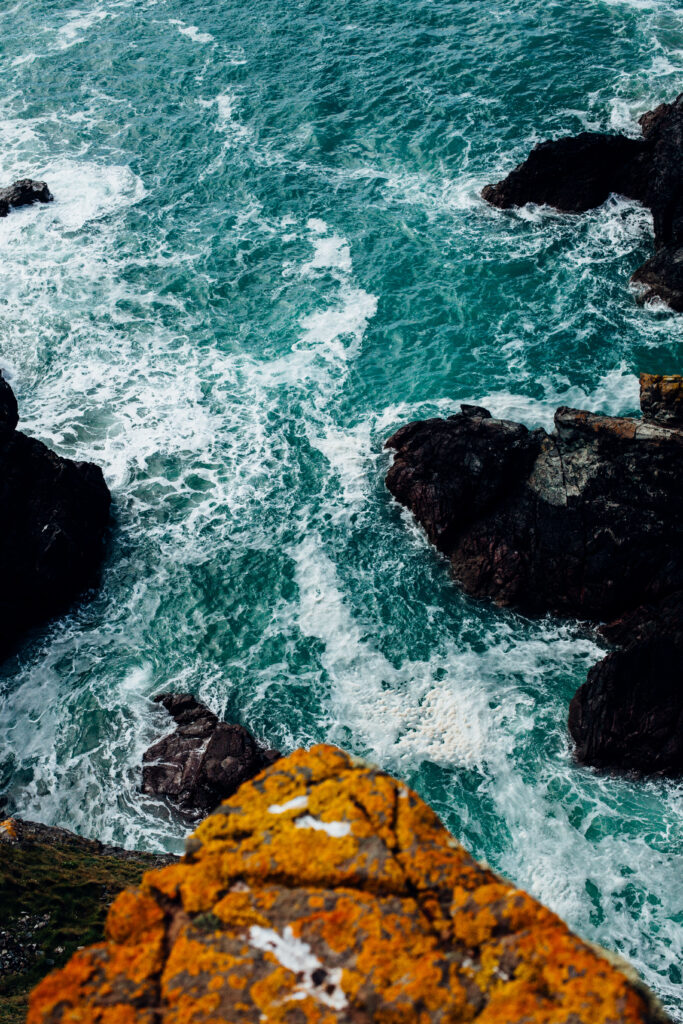 Turquoise waves crash on rugged rocks with orange lichen and white sea foam coastal wallpaper background