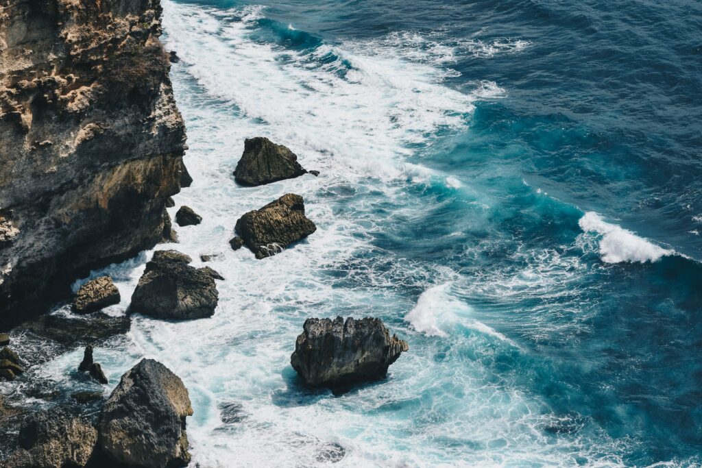 Turquoise sea waves crashing on rugged rocky coastline with foamy white water and textured rocks Wallpaper