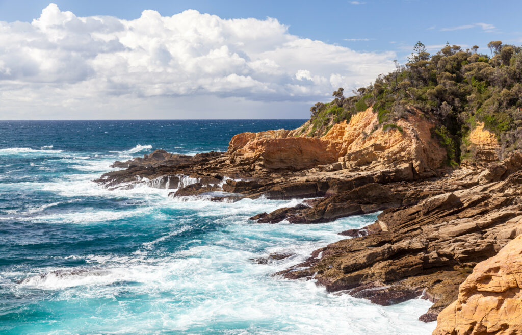 Turquoise waves crash on rugged stone cliffs with lush vegetation under a partly cloudy sky Wallpaper