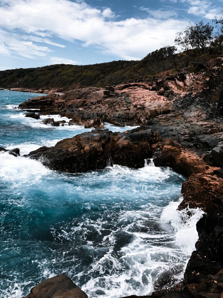 Dramatic turquoise waves crashing on rugged red rocks under partly cloudy sky coastal wallpaper