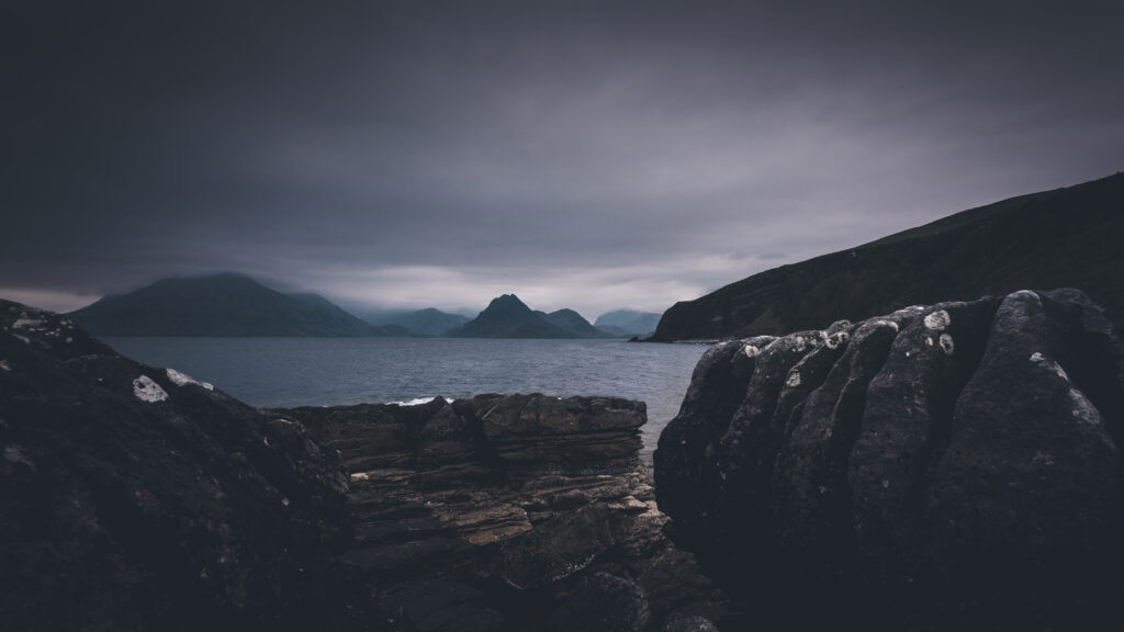 Dramatic rugged rocks by sea under twilight sky with distant mountains and moody clouds wallpaper