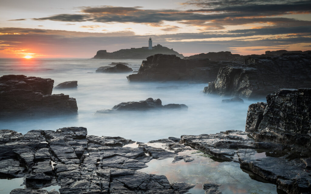 Twilight seascape with rugged rocks foggy shoreline and lighthouse silhouette at sunset wallpaper