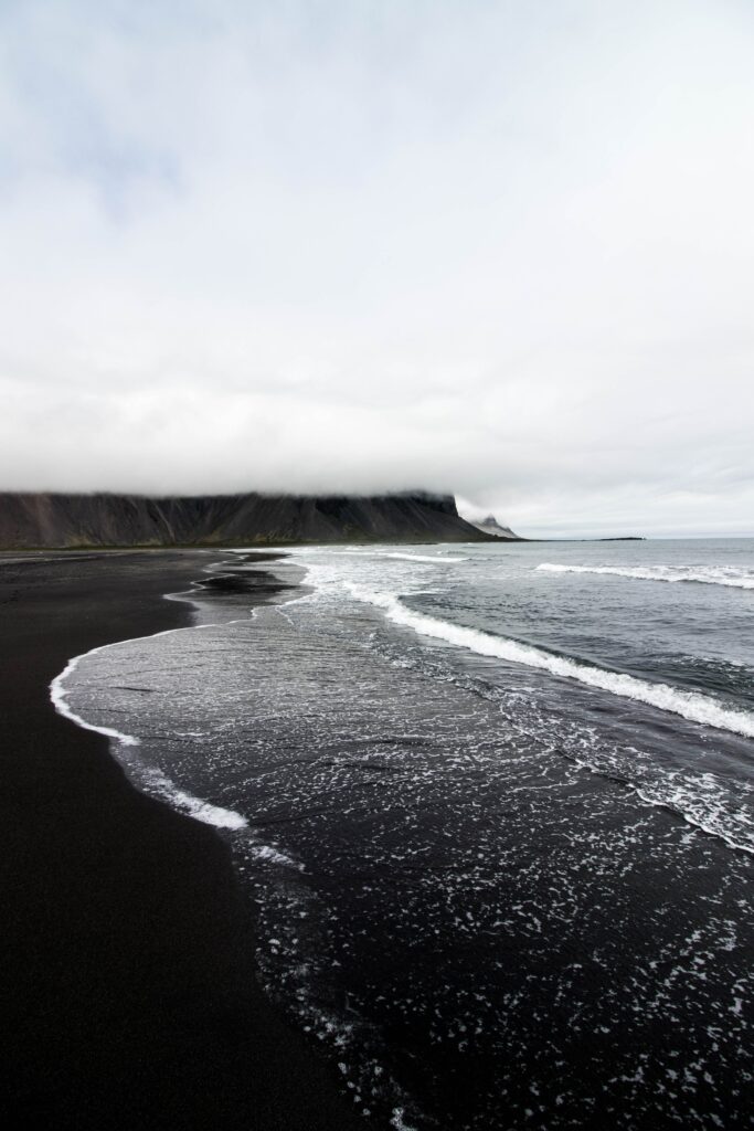 Rugged volcanic black sand beach with rolling waves misty clouds and distant cliffs wallpaper background