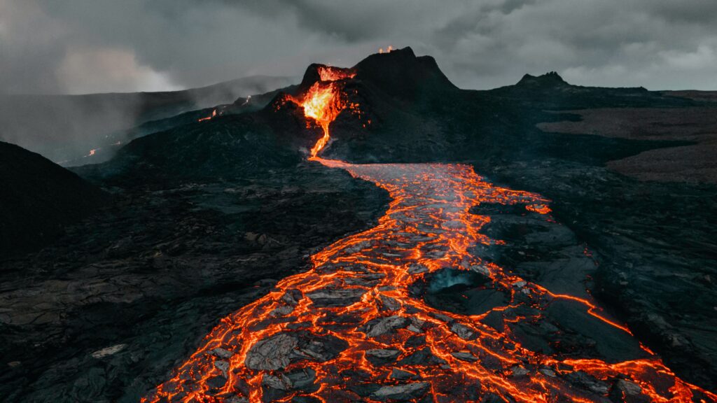 Dramatic volcanic eruption with bright molten lava flowing over black stones under stormy skies Wallpaper