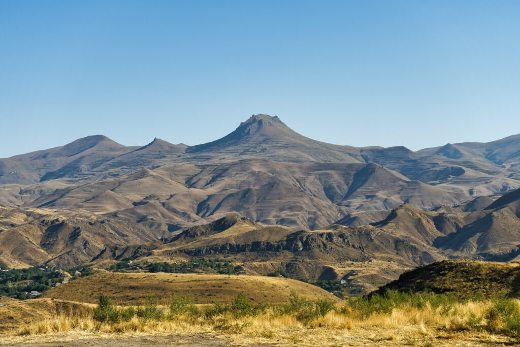 Volcanic mountain peak above layered rugged hills in warm earthy tones under clear blue sky Wallpaper