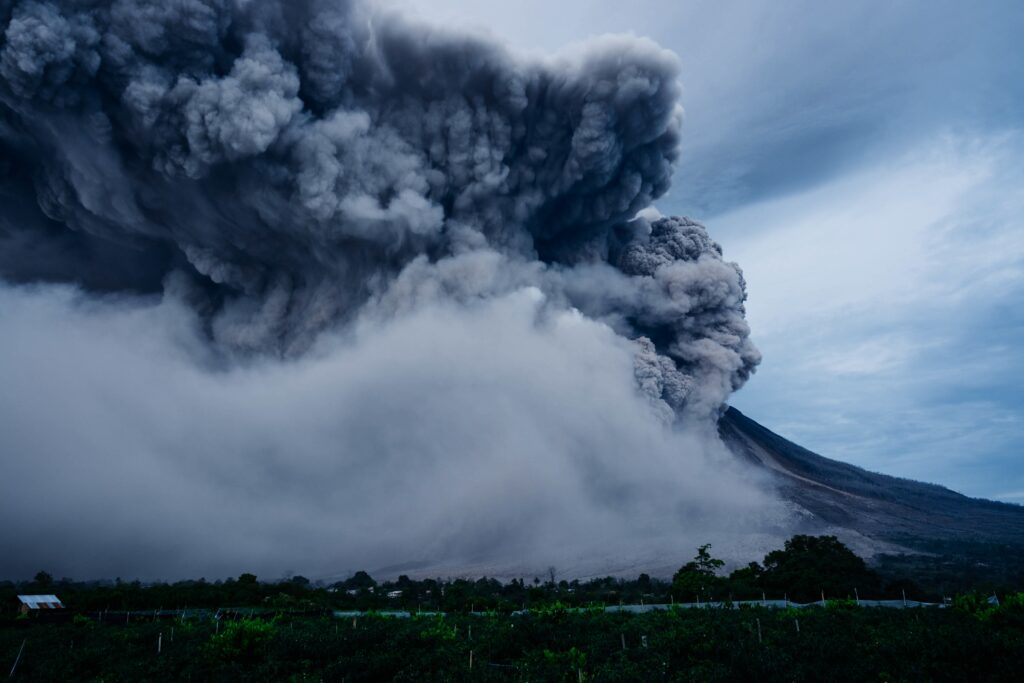 Erupting volcano with dark ash clouds over lush green landscape under moody overcast sky Wallpaper