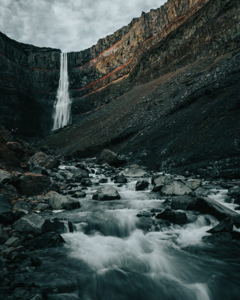 Dramatic waterfall cascading from rugged cliff into rocky stream with dark stones and moody colors Wallpaper