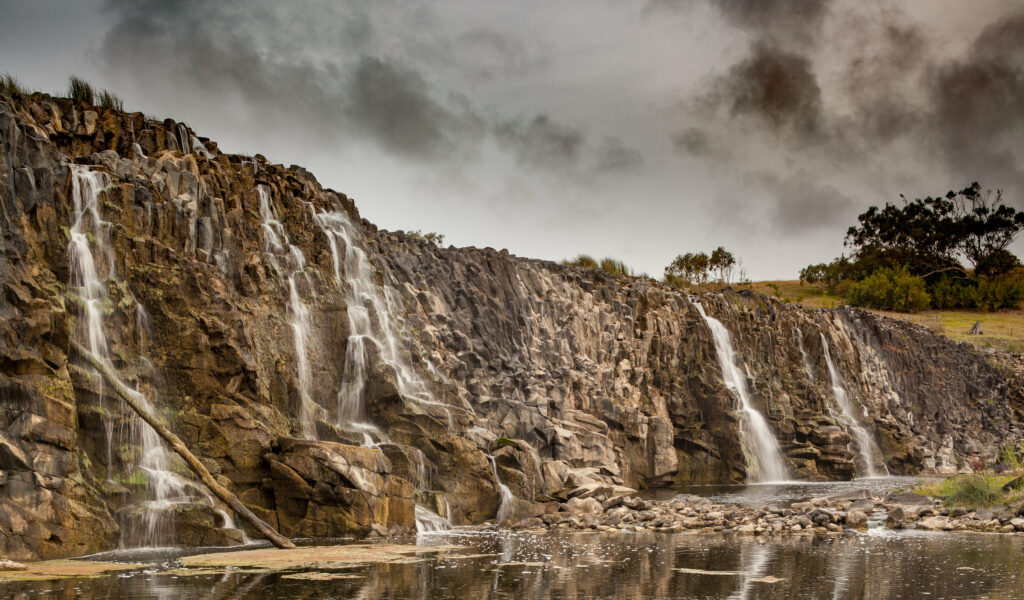 Dramatic waterfall cascading down jagged cliffs with sparse trees under cloudy moody sky wallpaper