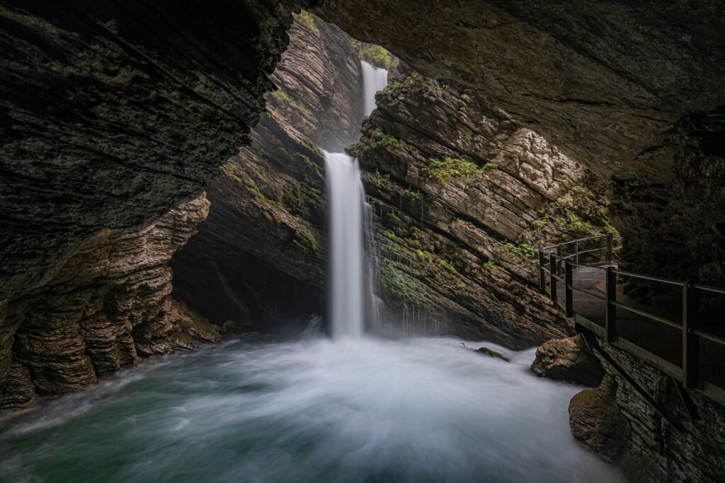 Dramatic waterfall cascading through rocky cave into misty blue stream with mossy greenery Wallpaper