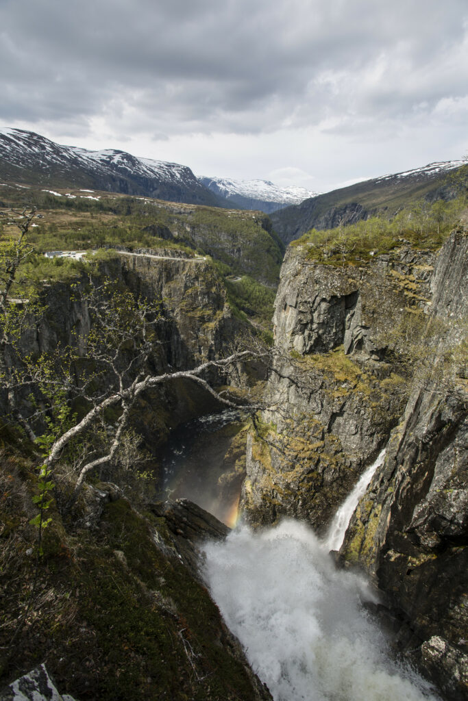 Dramatic cliffs and towering rocks frame a roaring river waterfall with snow-capped mountains background Wallpaper
