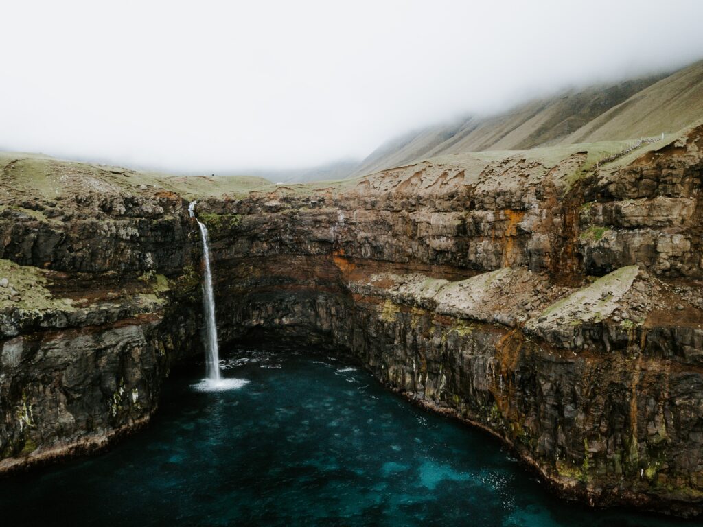 Dramatic waterfall from cliff into deep blue stream with rocky terrain and green grass landscape Wallpaper